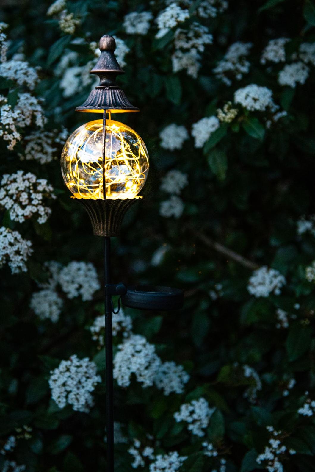 Decorative anthic-style solar lamp on a post with a ball containing a filament of small lights surrounded by a rust-colored pointed metal cap. Garden ID